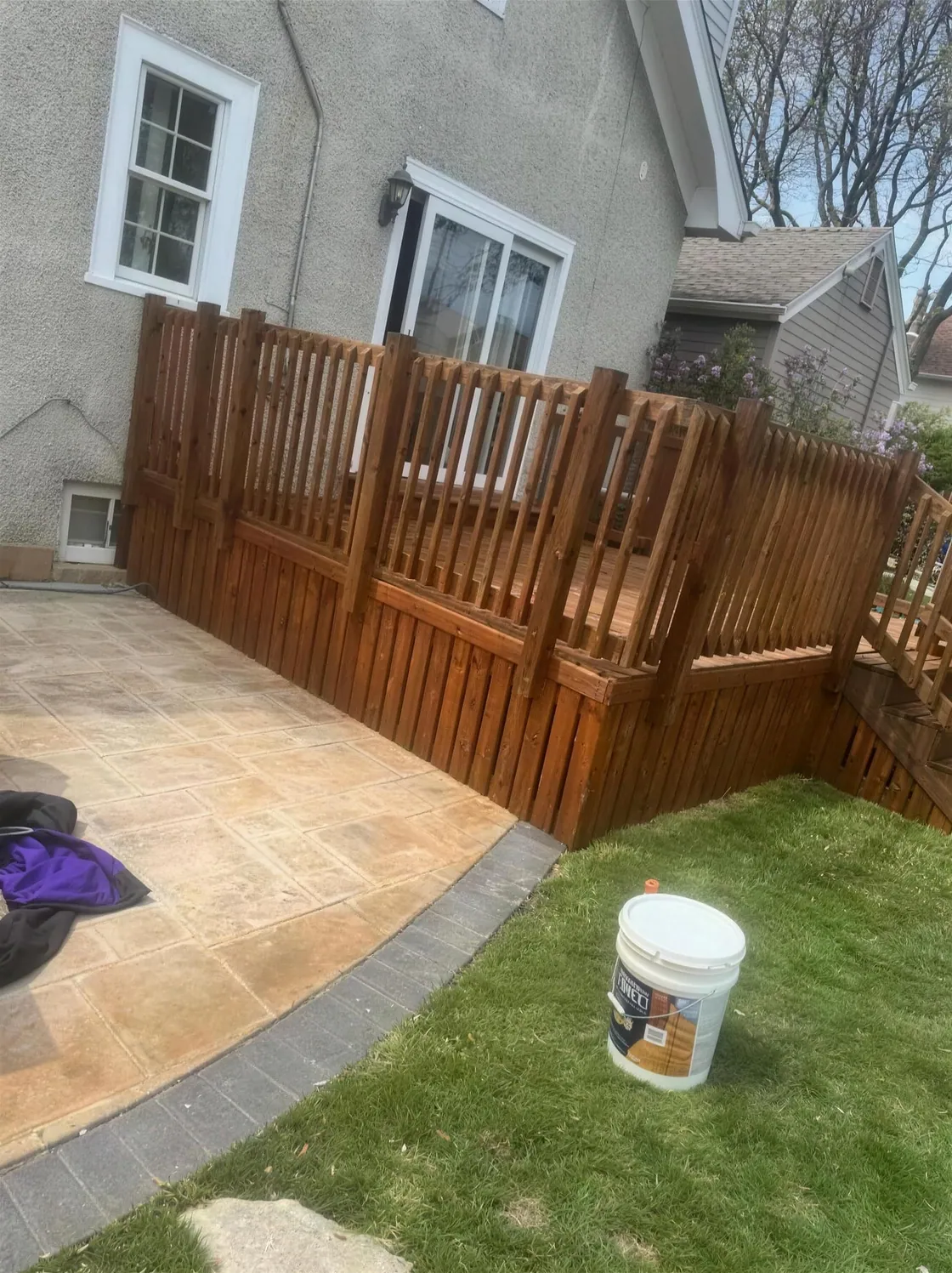 A wood deck and railing freshly stained a warm brown beside a stamped concrete patio, the stain bucket still on the lawn