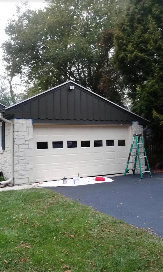 A detached garage part way through a repaint, the gable in black, the door and body in cream, a ladder and drop cloth still in place