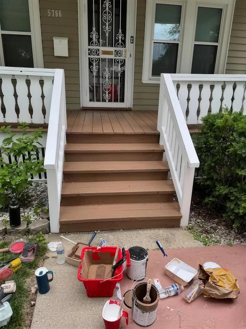 A front porch part way through a repaint, the steps and deck in fresh colour, the railings white, paint cans and buckets still on the walk